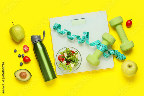 Фотография Weight scales with measuring tape, dumbbells, bottle of water and healthy food on yellow background