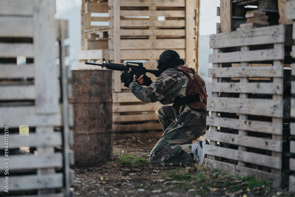Fototapeta premium A camouflaged player crouches behind pallets, aiming a paintball marker in a staged outdoor course.