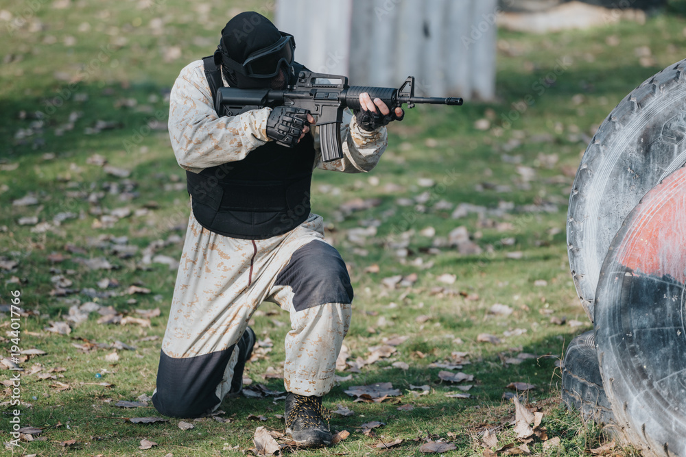 Fototapeta premium A person wearing a helmet, vest and camo kneels and aims a rifle on a paintball field. The scene shows a focused tactical player in protective gear amid an outdoor park setting.