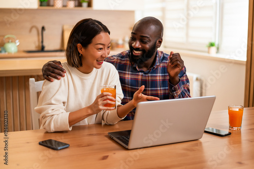 A joyful couple spends time together online, reacting happily to content on a laptop while enjoying drinks in a comfortable kitchen environment.