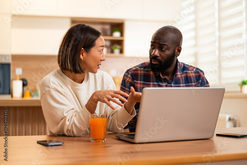 Adult couple sits together at a wooden table, using laptop for video communication in a bright and cozy home kitchen.