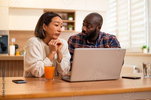 Adult couple sits together at a wooden table, using laptop for video communication in a bright and cozy home kitchen.