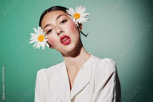 Creative beauty portrait of a young woman with bold makeup and daisy accessories touching her tongue playfully. Fashion photo with striking green background and white clothing symbolizes freshness.