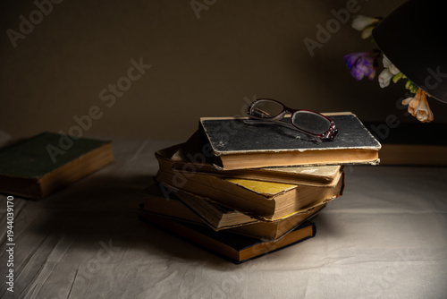 Stack of old books with reading glasses illuminated by desk lamp, cozy low light study still life.