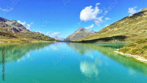 A tranquil lake mirrors rugged hills and towering mountains beneath bright skies. Green and brown terrain blends with clouds, creating a peaceful scene marked by vivid natural colors.  