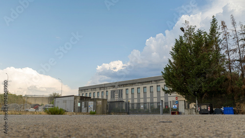 A wide shot of a modern industrial building under a bright blue sky with fluffy white clouds. Green trees frame the right side, while a gravel foreground leads to a gated entrance and utility sheds.
