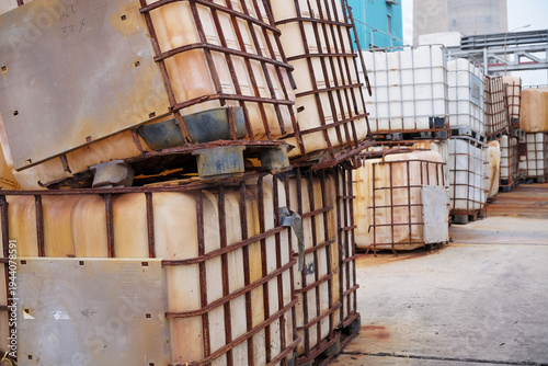 Industrial Storage Area Featuring Numerous Rusty Stacked Intermediate Bulk Containers Used for Chemical Waste Including Hcl 32 Percent Acid at an Outdoor Plant Facility