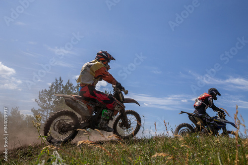 Two motocross riders racing side by side on grassy hillside dirt track, kicking up dust under