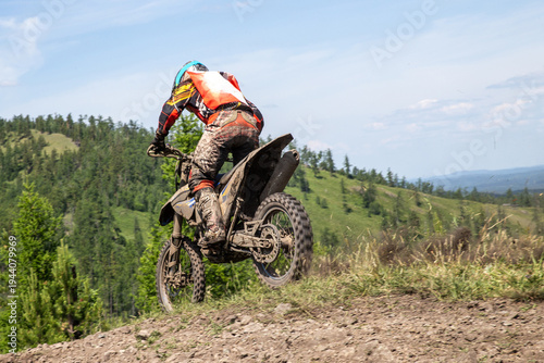 Rear view enduro rider on heavily mud-covered dirt bike performing wheelie on rocky hilltop trail