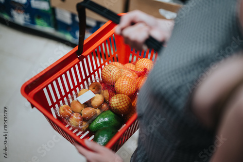 A woman holds a red shopping basket filled with oranges, avocados and packaged apricots. She shops for fresh fruit in a supermarket aisle, viewed from above.