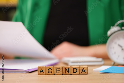 In a warm and inviting workspace, a person reviews documents while focusing on an agenda made of wooden blocks. An alarm clock adds to the sense of time management and productivity.