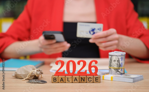 A person in a bright red jacket holds a phone and a credit card while arranging cash and a bag of coins. The year 2026 stands out, symbolizing future financial goals and balance.