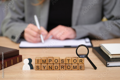 Bild auf Leinwand In a contemporary office, a person is focused on writing notes while blocks spelling out impostor syndrome sit nearby, illustrating the struggle with self-doubt and confidence