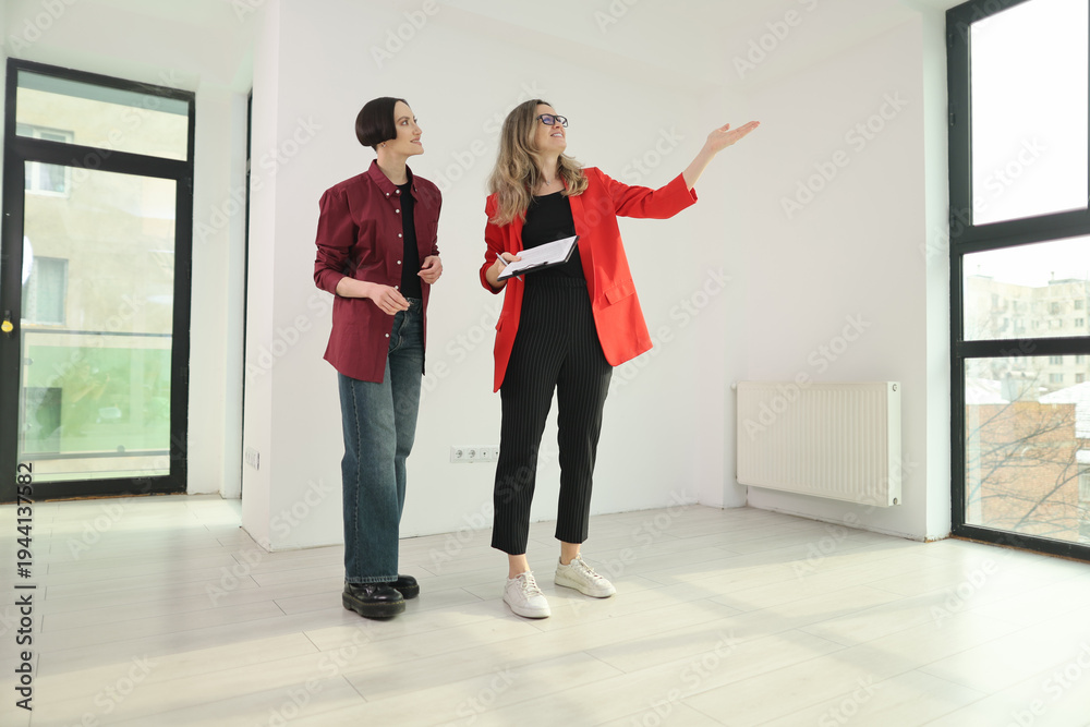 Fototapeta premium Two women stand in a bright, empty room filled with natural light, discussing design options. One gestures towards the window while holding a clipboard, inspiring creativity.