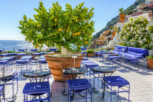Amalfi coast, Italy. Outdoor seating area featuring blue chairs and tables, a lemon tree in a pot, and a view of the sea and hillside buildings