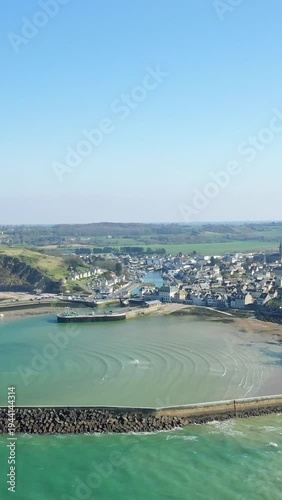 Scenic aerial shot of Port en Bessin harbor with turquoise waters, coastal village, and surrounding countryside under a clear blue sky in Normandy, France.