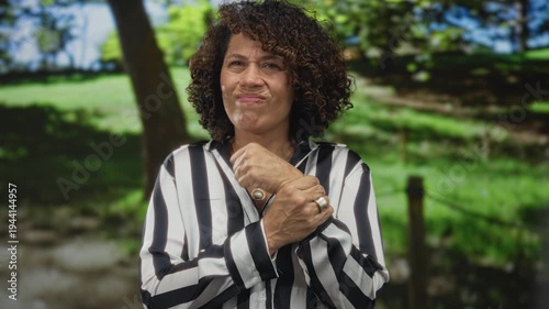 Hispanic middle aged woman in striped shirt grips wrist while grimacing in lush green park with blurred trees; pain injury discomfort.
