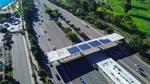 Toll plaza shoreline solar roofed roadway structure beside skatepark, beach edge, palm lined greenspace at Glorietta Bay. Lane shadows, bright concrete curves, warm coastal light, Coronado, CA