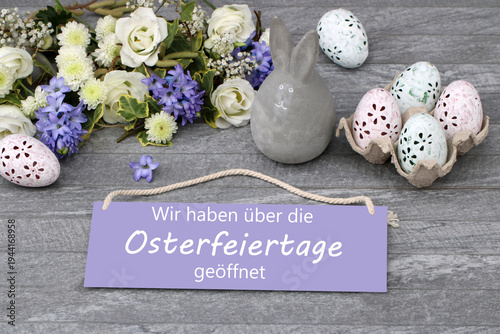 Öffnungszeiten an Ostern.: Schild mit der Aufschrift  Wir haben über die Osterfeiertage geöffnet mit Osterdekoration und Blumen.