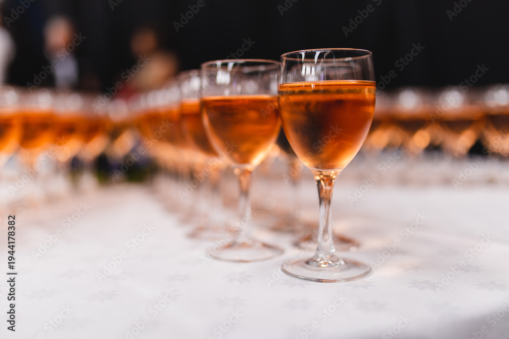 Fototapeta premium Rows of elegant wine glasses filled with chilled rose wine or amber cider on a white tablecloth. Shallow depth of field with a blurred background of a festive event, celebration, or gala dinner.