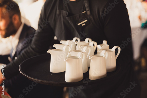 Professional waiter in a black apron carrying a round tray filled with several white ceramic milk pitchers or creamers, serving guests at a catered event, banquet, wedding, or elegant restaurant.