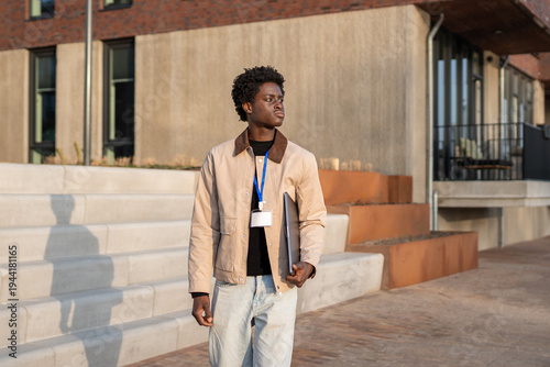 Photos Young black man carrying a laptop and id badge outdoors, facing right with a con