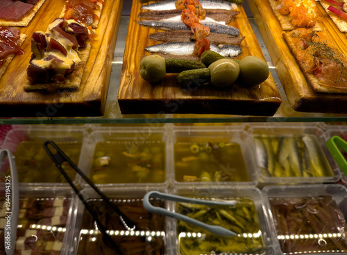 Selection of traditional Spanish tapas displayed on wooden boards inside a bar counter. Anchovies, salmon, tuna and various appetizers served with olives and pickled vegetables. Typical Mediterranean 