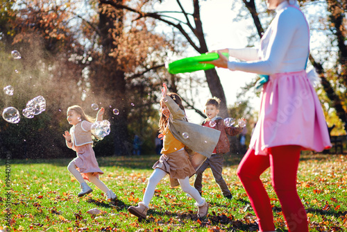 Female party animator playing with children at birthday party making soap bubbles while running in autumn park.