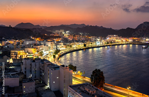 A stunning panoramic view of the illuminated Mutrah Corniche and harbor area at sunset, showcasing the coastal architecture and mountainous backdrop in Muscat, Oman.