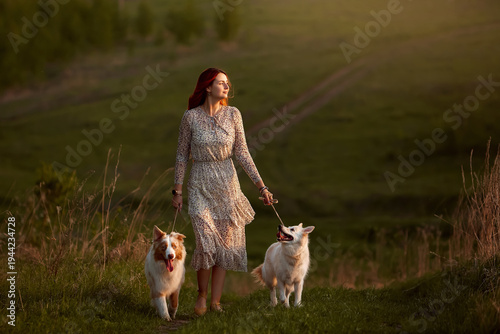 Young redhead woman in floral summer dress walking two dogs on green hill trail during golden hour sunset light. Romantic country lifestyle with pets in nature. Beautiful evening scene with copy space