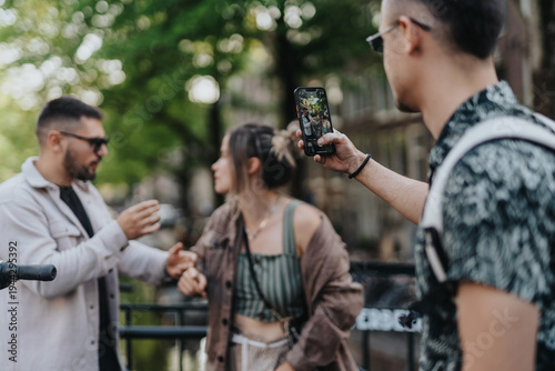 Three friends pose and chat while one man photographs them with a smartphone on a canal bridge. A candid street moment shows casual young people socializing in Amsterdam during summer.