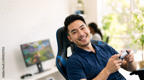 Portrait of a smiling Asian male gamer holding a white controller. Happy young man sitting in a gaming chair playing video games in a bright room