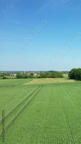 Wide view of lush green fields under a clear blue sky near the Hillman Batteries in Normandy, France. Trees and distant rural buildings complete the peaceful countryside scene.
