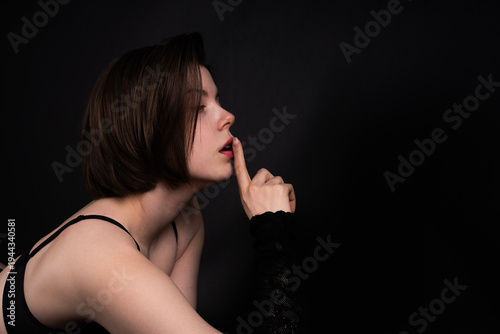 Foto Woman in lingerie lying on stomach on black background making hush gesture with finger on lips, erotic concept, side profile, low key studio lighting