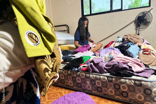 Young Filipino woman in bedroom sorting and folding large pile of clean laundry after washing, Asian lady organizing clothes on bare mattress at home