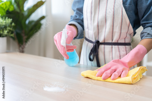 Fotografi Cleanliness asian young woman working chore cleaning on table at home, hand wearing glove using rag rub remove dust with spray bottle