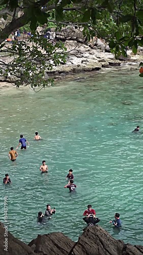 People swimming and playing in clear turquoise water near a rocky shoreline with trees.