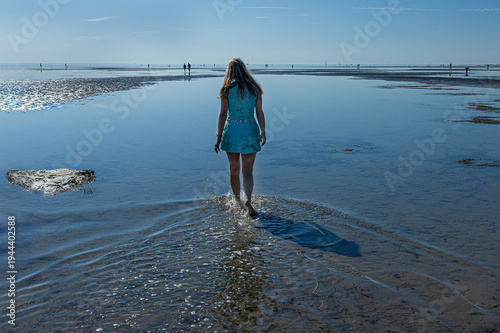 Woman walking on wide tidal flats with beautiful ripple patterns in the sand during low tide. Minimalist coastal landscape with calm sea and clear blue sky.