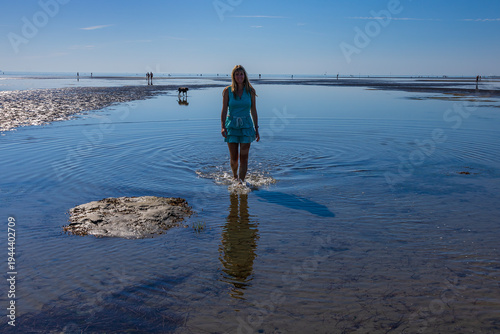 Woman walking on wide tidal flats with beautiful ripple patterns in the sand during low tide. Minimalist coastal landscape with calm sea and clear blue sky.