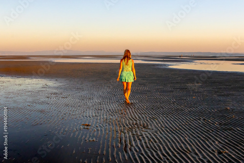 Woman walking on wide tidal flats with beautiful ripple patterns in the sand during low tide. Minimalist coastal landscape with calm sea and clear blue sky.