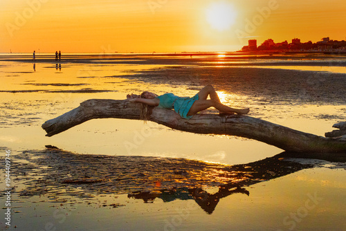Woman lying on a driftwood log on wide tidal flats with ripple patterns in the sand at low tide. Minimalist coastal landscape with calm sea and setting sun in the background.