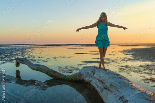 A woman walks along driftwood on a wide tidal flat with beautiful wave patterns in the sand at low tide. Minimalist coastal landscape with a calm sea and the setting sun in the background.