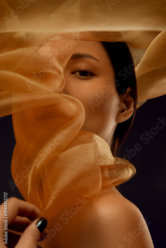 Beauty portrait of Asian woman framed by translucent golden silk fabric with flowing shapes for premium skincare advertising, luxury cosmetics branding and beauty marketing.