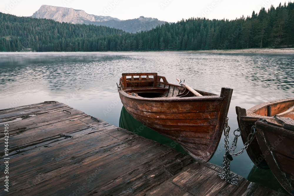 Obraz premium Wooden boat moored at a pier on Black Lake in Durmitor National Park, Montenegro, with calm water, forest and mountain backdrop, ideal for travel and nature themes.