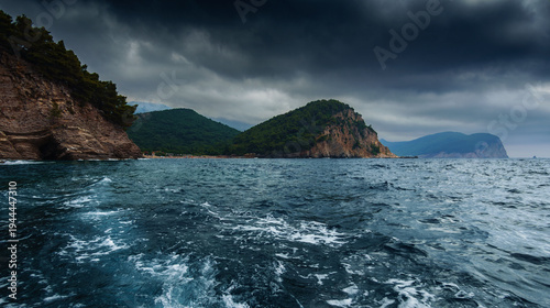 View from a boat of the rugged Petrovac coastline with mountains and splashing sea wake under dark clouds.