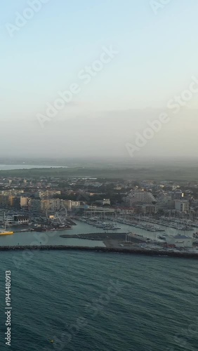 Scenic aerial shot of the marina and waterfront in Canet-en-Roussillon, France, with boats, buildings, and a Ferris wheel at sunset.
