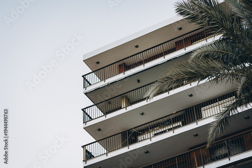 Low angle view of modern apartment building balconies with metal railings and palm tree fronds, captured against a clear sky for urban living, travel, and architecture concepts.