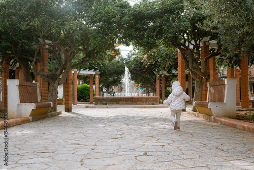 Rear view of small child in warm clothes walking alone along stone path toward water fountain in peaceful urban park with benches, trees and brick columns.