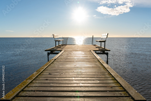 фотографія A straight wooden pier leads toward open water and a bright sunlit horizon, with simple benches at the end