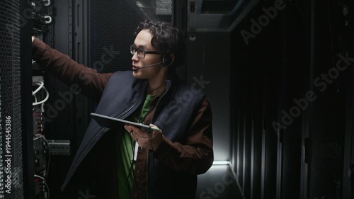 Indoor medium slowmo shot of Asian male network technician standing by opened server rack checking cables and ports while reviewing on tablet and communicating via headset in dark data center